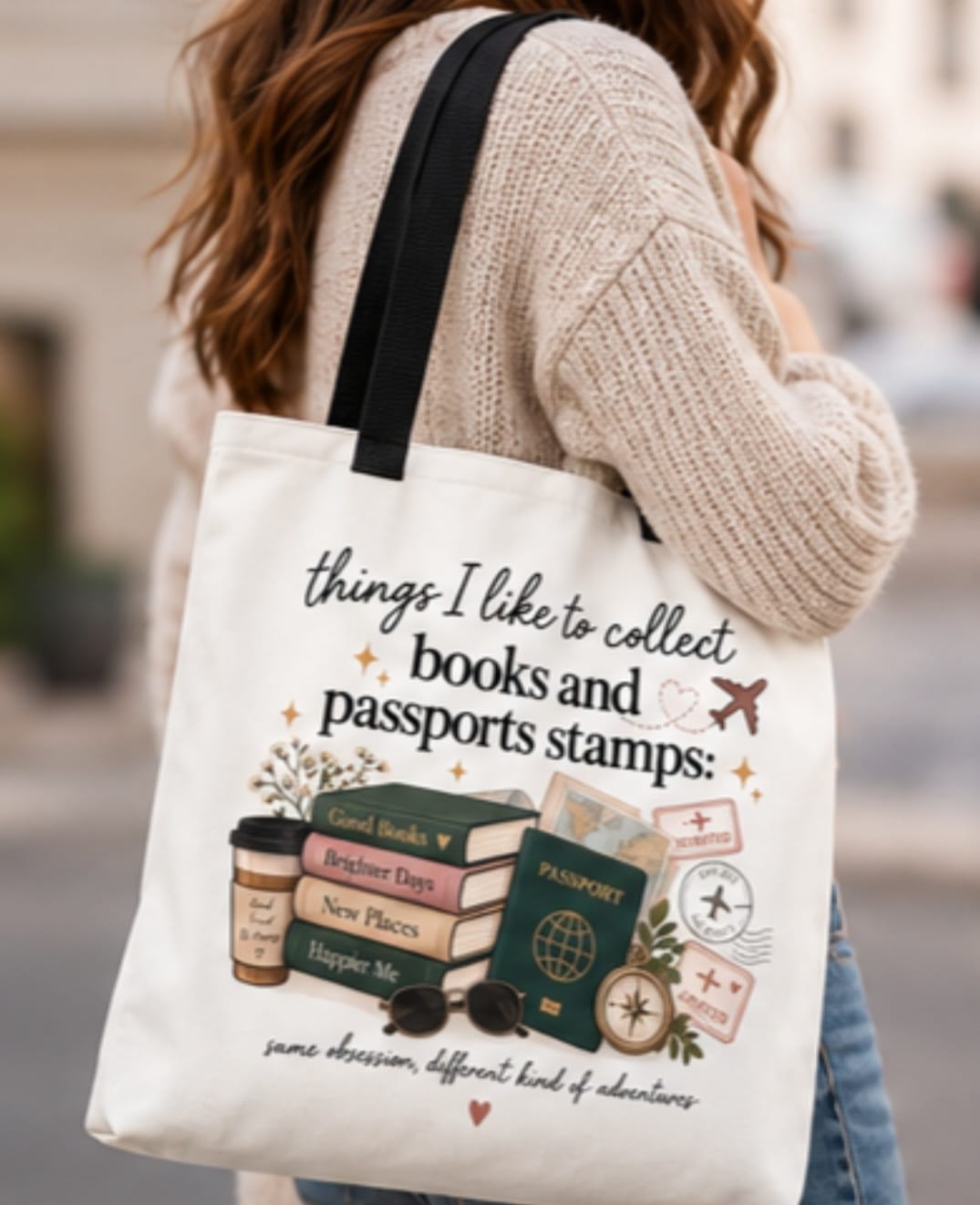 White canvas tote bag with black handles, printed passport stamps and book-themed illustration